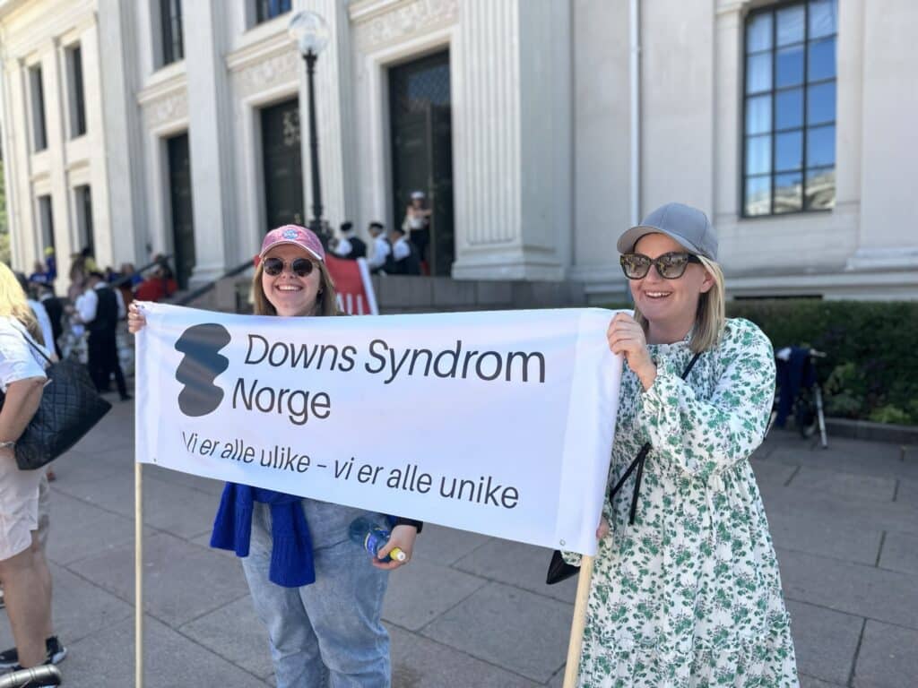 To damer som holder et banner med teksten Downs Syndrom Norge - vi er alle ulike, vi er alle unike