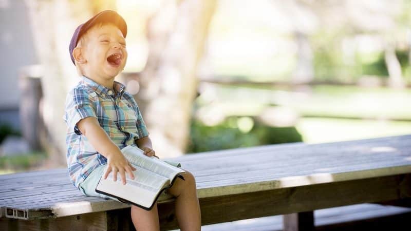 Gutt som sitter på en benk med en bok på fanget og ler